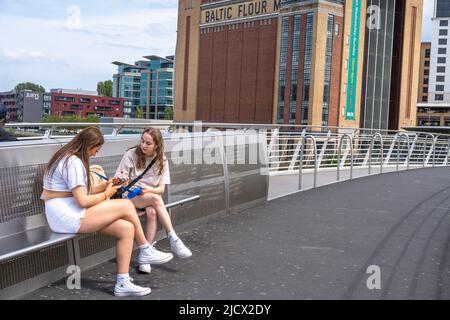 Deux jeunes femmes sont assises sur le Gateshead Millennium Bridge à Newcastle upon Tyne, Royaume-Uni avec la galerie d'art BALTE à Gateshead derrière. Banque D'Images