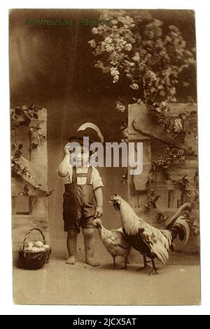 Carte postale originale kitsch Austrian publiée carte de vœux de Pâques destinée au marché français, représentant un joli petit garçon portant un short traditionnel de style lederhosen (short en cuir), un chapeau avec plumes, poules et maquereau, panier d'œufs, Heuruses Paques (Happy Easter) écrit sur le devant, publié par W.R.B. & Co. Vienne, en poste le 11 avril 1914 depuis Paris, juste avant le début de l'épidémie de WW1. Banque D'Images