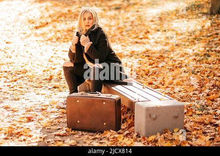 L'automne est l'heure du changement. Beau blond féminin en manteau de cachemire noir et jeans est assis sur le banc de jardin dans le parc de la ville, vieille valise est Banque D'Images