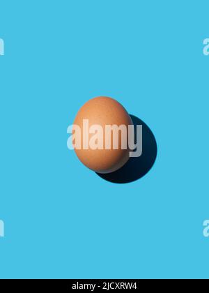 Un œuf de poulet brun se trouve au centre sur un fond bleu vif. Lumière vive, ombre profonde, verticale. Gros plan, espace de copie, vue de dessus. Banque D'Images