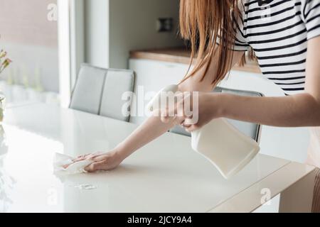 Photo rognée de la jeune femme se tenir dans la cuisine pulvérisation sur table blanche du pulvérisateur, essuyage avec un chiffon, désinfection. Banque D'Images