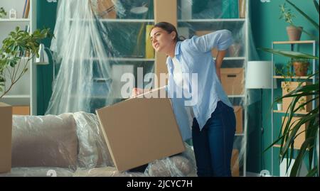 Une femme ressent une douleur soudaine au dos lorsqu'elle tente de soulever une boîte à carton. Femme utilisant un massage de la main sur sa douleur de dos. Une femme éprouve une gêne dans un résultat Banque D'Images