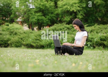 Jeune femme assise sur l'herbe verte dans un parc avec les jambes croisées un jour d'été tout en utilisant un ordinateur portable Banque D'Images