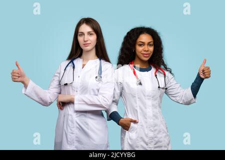 Sympathique deux femmes médecins GP montre le pouce vers le haut, geste d'approbation, le signe OK, comme sur l'arrière-plan de Blue Studio. Groupe multiethnique substance médicale de Healthc Banque D'Images