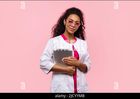 Femme médecin médecin médecin à la peau foncée tenant une tablette numérique avec antécédents médicaux dans un dossier clinique électronique sur un mur de studio rose. Femme Med Banque D'Images