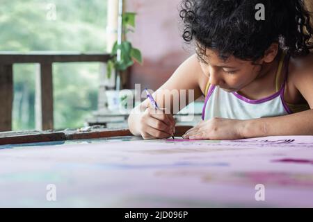 pauvre latina fille avec la peau brune très concentré peindre son dessin. elle a fait la vaisselle avec les cheveux frizzy et les vieux vêtements faisant ses devoirs à un bureau Banque D'Images