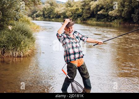 Pêcheur à la ligne passionné homme senior pêcheur dans chapeau de cowboy avec canne à pêche, tournant bobine sur la rivière. Vieil homme attrapant du poisson, tirant la tige tout en pêchant sur le lac. Banque D'Images