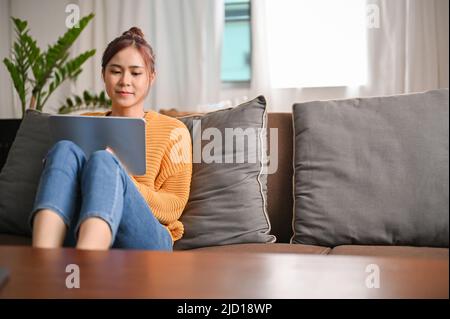 Une jolie jeune femme asiatique passe son week-end à la maison, assise sur le confortable canapé dans le salon et utilisant une tablette numérique portable. Banque D'Images