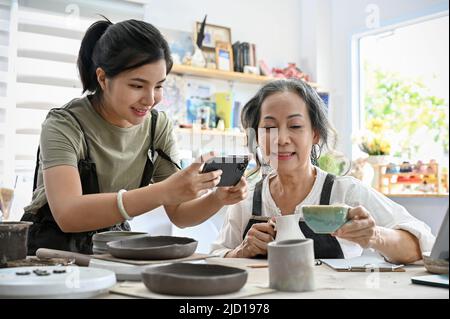 Jeune femme asiatique prenant une photo d'une tasse de café en céramique dans les mains d'une femme âgée dans l'atelier d'artisanat. Banque D'Images