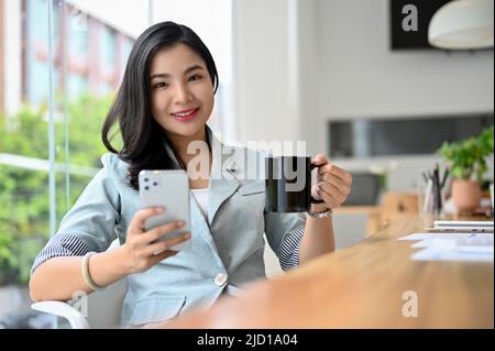 Charmante jeune femme d'affaires asiatique ou femme de direction qui sirotait un café tout en utilisant un smartphone à son bureau moderne. Banque D'Images