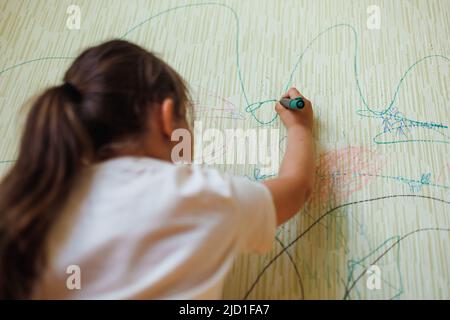 Vue arrière de la petite mauvaise fille avec cheveux foncés tenant le marqueur vert à la main, dessin d'images sur le vieux papier peint jaune. Banque D'Images