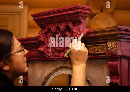 Femme attentive colorant un placard en bois massif avec des ornements sculptés étonnants en rouge avec une brosse avec des murs de maison en arrière-plan. Réutilisation de l'ancien Banque D'Images