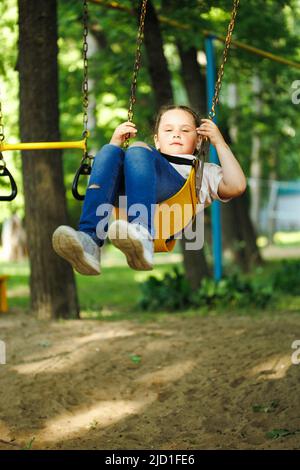 Une petite fille mignonne qui fait du thé sur une balançoire jaune dans un terrain de jeu dans un parc verdoyant. Vacances d'été dans le camp, centre touristique. Marcher et jouer à l'extérieur, sport Banque D'Images