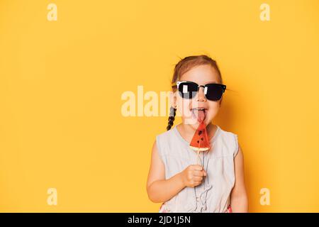 Fille en lunettes licks lollipop sous forme de morceau de pastèque sur fond jaune Banque D'Images