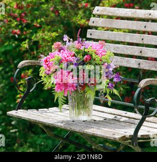 Bouquet coloré dans les tons rouge, rose et violet avec des pivoines et des colonnes, supports en vase de verre sur banc de jardin décoratif en bois Banque D'Images