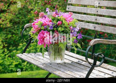 Bouquet coloré dans les tons rouge, rose et violet avec des pivoines et des colonnes, supports en vase de verre sur banc de jardin décoratif en bois Banque D'Images