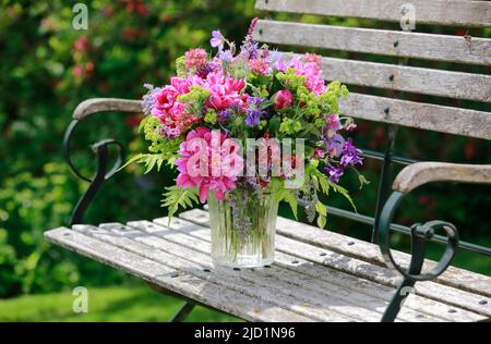 Bouquet coloré dans les tons rouge, rose et violet avec des pivoines et des colonnes, supports en vase de verre sur banc de jardin décoratif en bois Banque D'Images