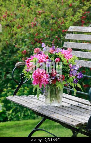 Bouquet coloré dans les tons rouge, rose et violet avec des pivoines et des colonnes, supports en vase de verre sur banc de jardin décoratif en bois Banque D'Images