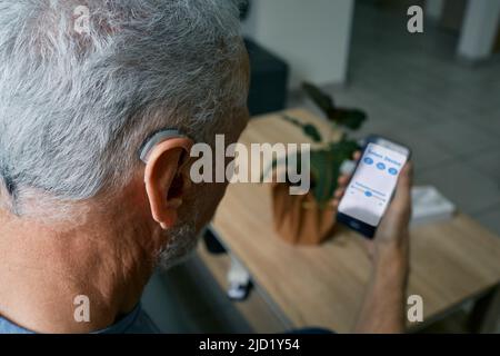 Homme senior malentendant qui règle les paramètres de son aide auditive BTE via un smartphone. Prothèses auditives avec technologies innovantes en audiologie Banque D'Images