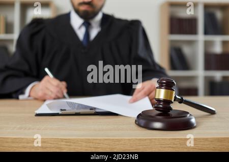 Gavel sur une table en bois dans un tribunal, avec un juge signant certains documents en arrière-plan Banque D'Images