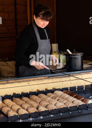 Karlovy Vary, Bohême, République Tchèque - 28 mai 2022 : une femme qui cuit le gâteau traditionnel de trdelnik au feu de charbon. Banque D'Images