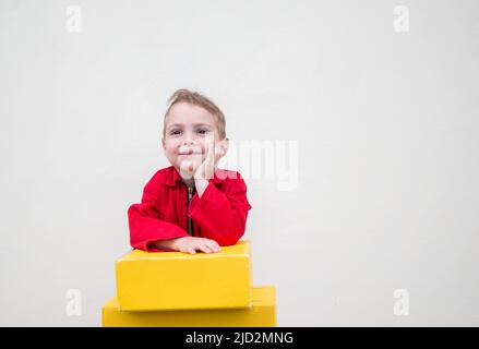 3-4-le visage heureux d'un garçon dans des combinaisons rouges près des boîtes en carton jaune pour les colis. Livraison, petit postier. Travaillez comme un papa. Commentaires positifs Banque D'Images