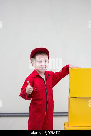 garçon de 3-4 ans dans une combinaison rouge, un uniforme de travail et un chapeau montre un signe similaire près des boîtes en carton jaune. atmosphère positive. livraison de bonheur. Po Banque D'Images