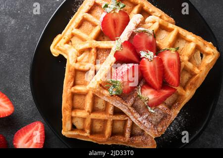 Gaufres belges. Gaufres maison avec fraises, sucre en poudre et tasse de café sur une assiette noire sur fond de table en pierre noire. Petit déjeuner. Haut v Banque D'Images
