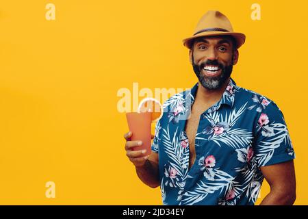 Beau barbu homme afro-américain adulte moyen portant une chemise hawaïenne et un chapeau souriant avec un cocktail de jus d'orange regardant le studio d'appareil photo Banque D'Images
