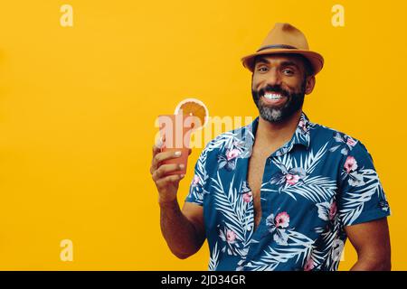 Beau barbu homme afro-américain adulte moyen portant une chemise hawaïenne et un chapeau souriant avec un cocktail de jus d'orange regardant le studio d'appareil photo Banque D'Images