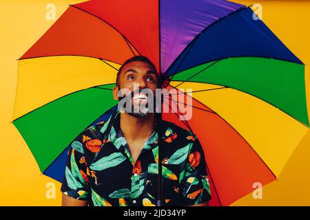 Beau sourire barbu africain américain dans une chemise hawaïenne posant avec un parapluie arc-en-ciel coloré en studio tourné Banque D'Images