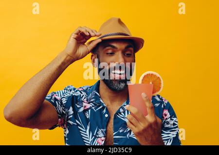 Homme afro-américain barbu adulte de taille moyenne portant une chemise hawaïenne et un chapeau regardant un studio souriant de jus d'orange Banque D'Images