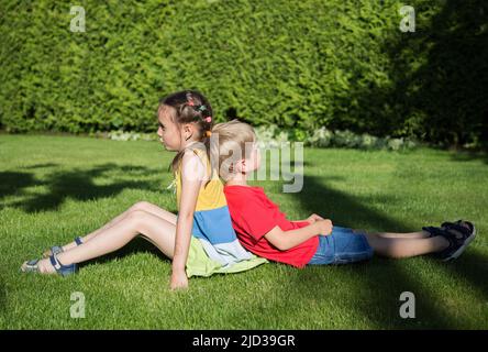 garçon et une fille de 6-7 ans sont assis dos à dos sur l'herbe dans le jardin. parler et jouer dans le parc vert ensoleillé. Amitié, enfance, rel Banque D'Images