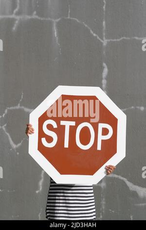 La personne porte le panneau de signalisation indiquant un grand arrêt. Rouge vif contre les murs gris fissurés. Symbole accrocheur pour aborder les concepts abstraits : comportement toxique et négativité. Banque D'Images