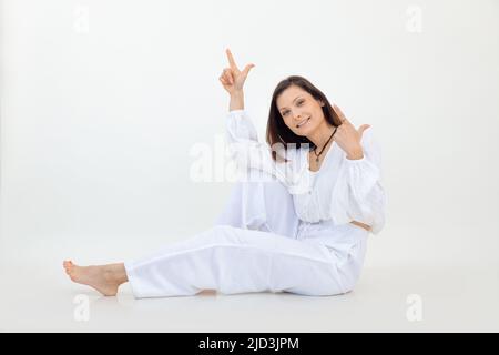 Femme souriante brune posant et assise sur un sol blanc de studio, regardant la caméra. Indication à l'aide des doigts. Copier l'espace Banque D'Images