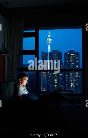 Guy se repose dans un canapé confortable avec ordinateur portable contre la fenêtre avec un paysage urbain époustouflant. Kuala Lumpur, Malaisie - 04.01.2020 Banque D'Images