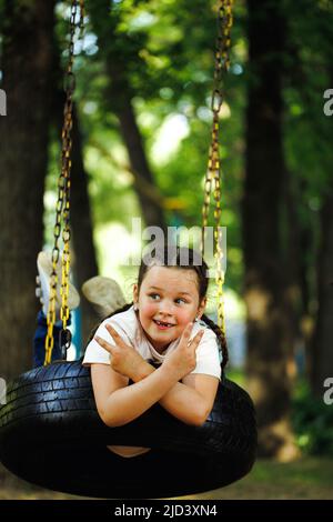 Petite fille fraîche balançant sur roue en caoutchouc dans l'aire de jeux dans parc vert de près. Vacances d'été dans le camp, centre touristique. Marcher et jouer à l'extérieur Banque D'Images