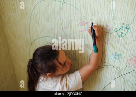 Vue de dessus de petite fille méchante avec cheveux foncés tenant le marqueur vert à la main, dessin d'images sur le vieux papier peint jaune. Banque D'Images