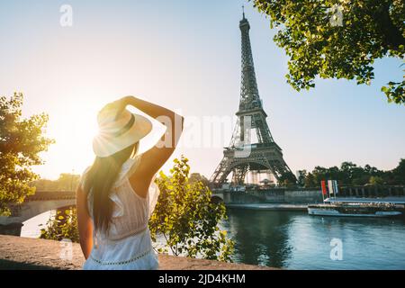 Jeune femme touriste en chapeau de soleil et robe blanche debout devant la Tour Eiffel à Paris au coucher du soleil. Voyage en France, concept de tourisme Banque D'Images