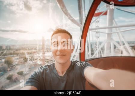 Un jeune homme heureux et excité prenant une photo de selfie en cabine d'une roue de ferris. Aventures de voyage solo et parc d'attractions Banque D'Images