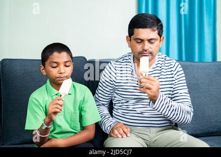 Le père et le fils s'affairent à manger des glaces à la maison tout en étant assis sur un canapé - concept d'été, de famille et de togethness. Banque D'Images