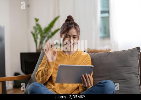 Jeune femme asiatique souriante dans un chandail jaune avec un ordinateur tablette PC pour passer un appel vidéo et passer la main à la maison Banque D'Images