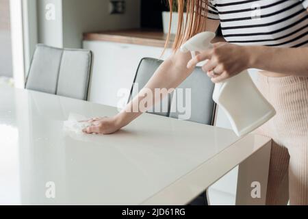 Femme méconnaissable se tenir dans la cuisine vaporisant sur une table blanche à partir d'un pulvérisateur en plastique, essuyant avec un chiffon, désinfectant. Banque D'Images