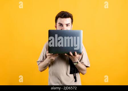 Beau jeune homme portant des vêtements décontractés posant isolé sur fond jaune regardant derrière l'ordinateur portable, surpris de la caméra. Banque D'Images