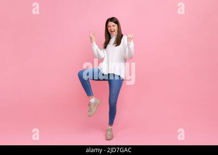 Portrait complet d'une femme excitée debout sur une jambe, montrant le mouvement de rock et de roulis, hurlant, portant blanc style décontracté pull. Studio d'intérieur isolé sur fond rose. Banque D'Images