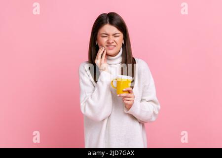 Malade brune femme souffrant de dents sensibles après avoir bu chaud ou froid boisson, cavités, portant blanc décontracté style pull. Studio d'intérieur isolé sur fond rose. Banque D'Images