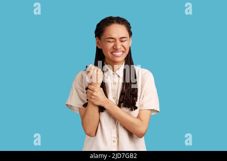 Contrarié mécontent femme avec des dreadlocks touche la main blessée, montre la zone douloureuse problématique sur le poignet, souffre de douleur terrible, portant une chemise blanche. Studio d'intérieur isolé sur fond bleu. Banque D'Images