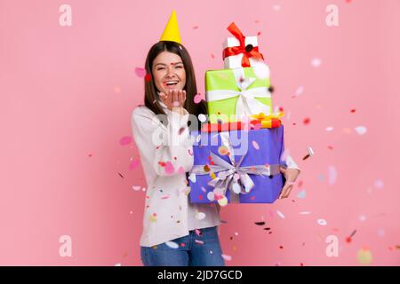 Femme extrêmement heureuse célébrant l'anniversaire tenant une pile de cadeaux, debout sous la pluie de paillettes, portant blanc décontracté style pull. Studio d'intérieur isolé sur fond rose. Banque D'Images