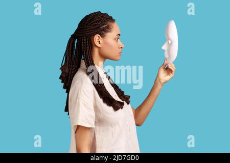 Vue latérale d'une femme sûre et sérieuse avec des dreadlocks tenant et regardant le masque blanc avec un regard attentif, essayant de comprendre la personnalité cachée. Studio d'intérieur isolé sur fond bleu. Banque D'Images