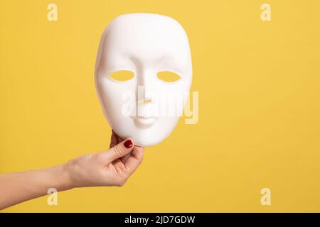 Profil vue de côté gros plan de la main de femme tenant le masque blanc modèle, masquant la personnalité. Studio d'intérieur isolé sur fond jaune. Banque D'Images